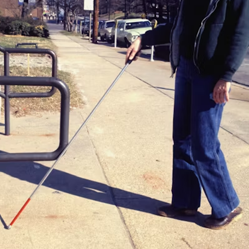 Person using a white cane while walking along a city sidewalk